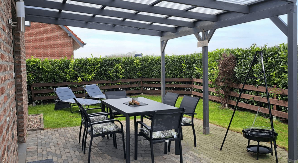 Ferienhaus Maas, überdachte Terrasse mit Tisch und Stühlen und Grill von Rasenfläche und Hecke umgeben