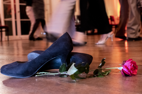 Damen-Tanzschuhe mit roter Rose auf Parkettboden mit Beinen/Schuhen tanzender Paare im Hintergrund