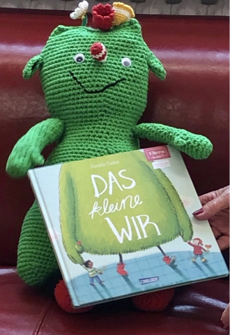 Zu sehen ist ein grünes Maskottchen mit einem Kinderbuch.